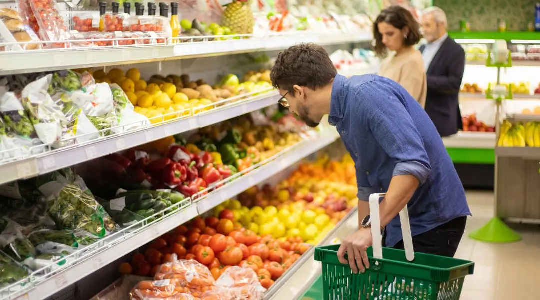 Personne en chemise bleue tenant un panier vert dans un rayon de supermarché rempli de fruits et légumes colorés