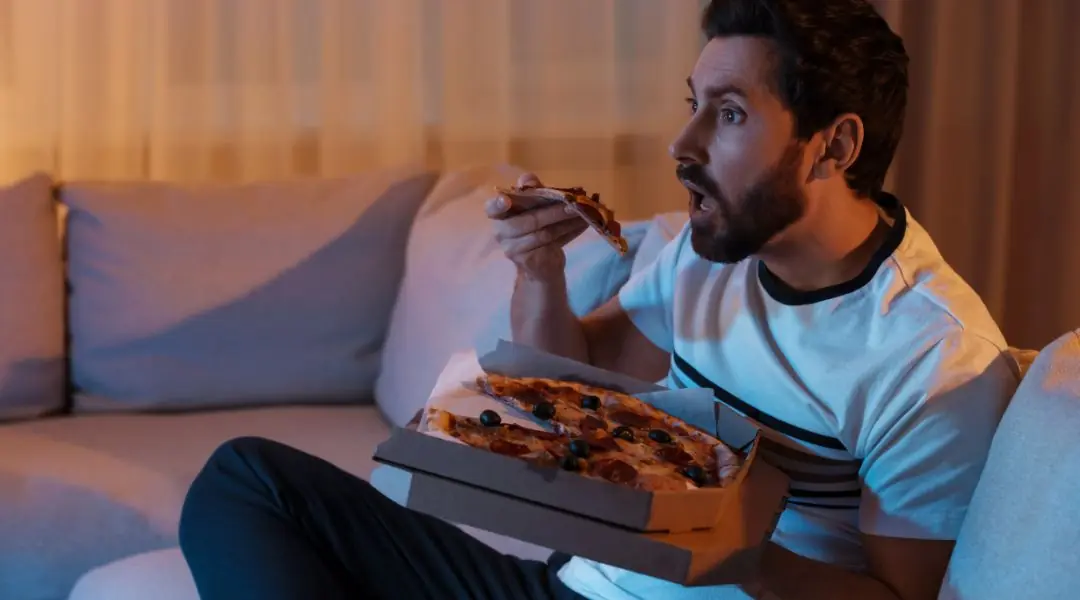 Person sitting on a couch, eating a slice of pizza from an open box in a dimly lit room