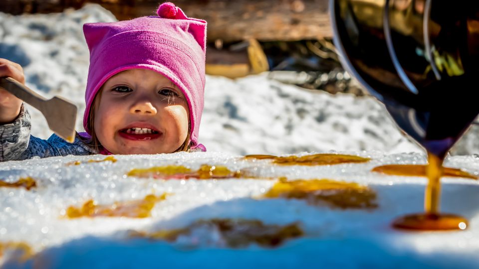 Un enfant souriant avec un bonnet rose déguste de la tire d’érable sur neige tandis que du sirop est versé