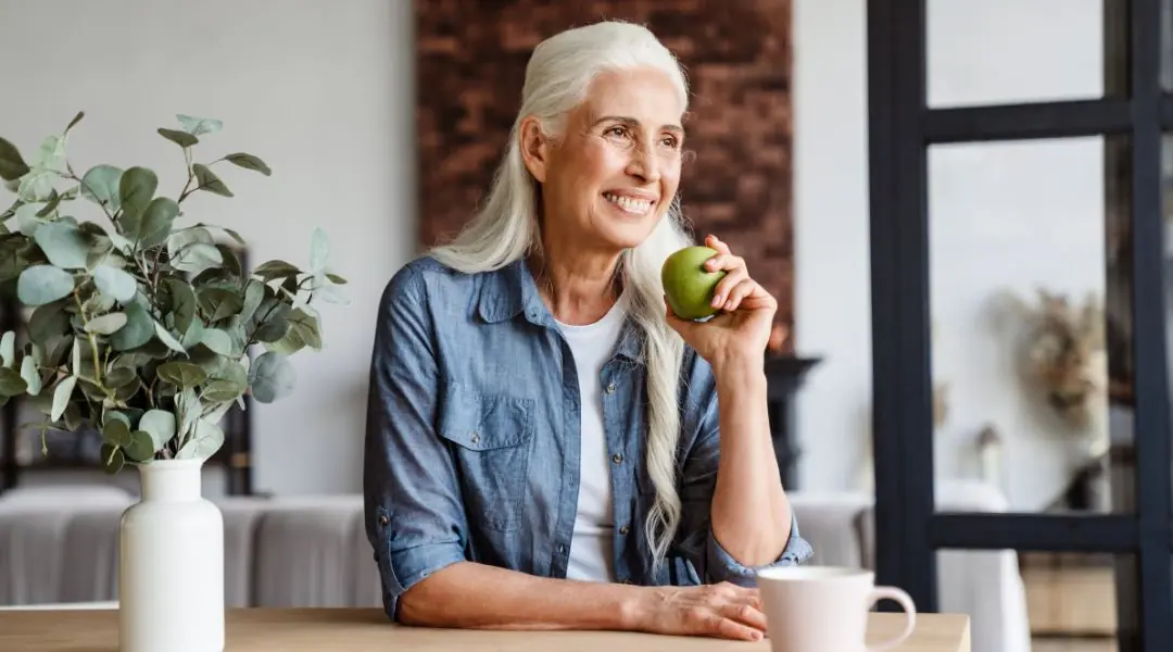 Femme aux cheveux blancs tenant une pomme verte, assise à une table avec une tasse et un vase de plantes