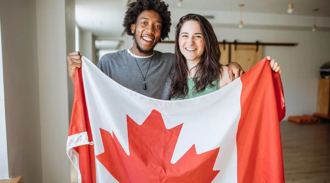 Deux personnes souriantes tenant un drapeau canadien dans une pièce lumineuse avec un sol en bois