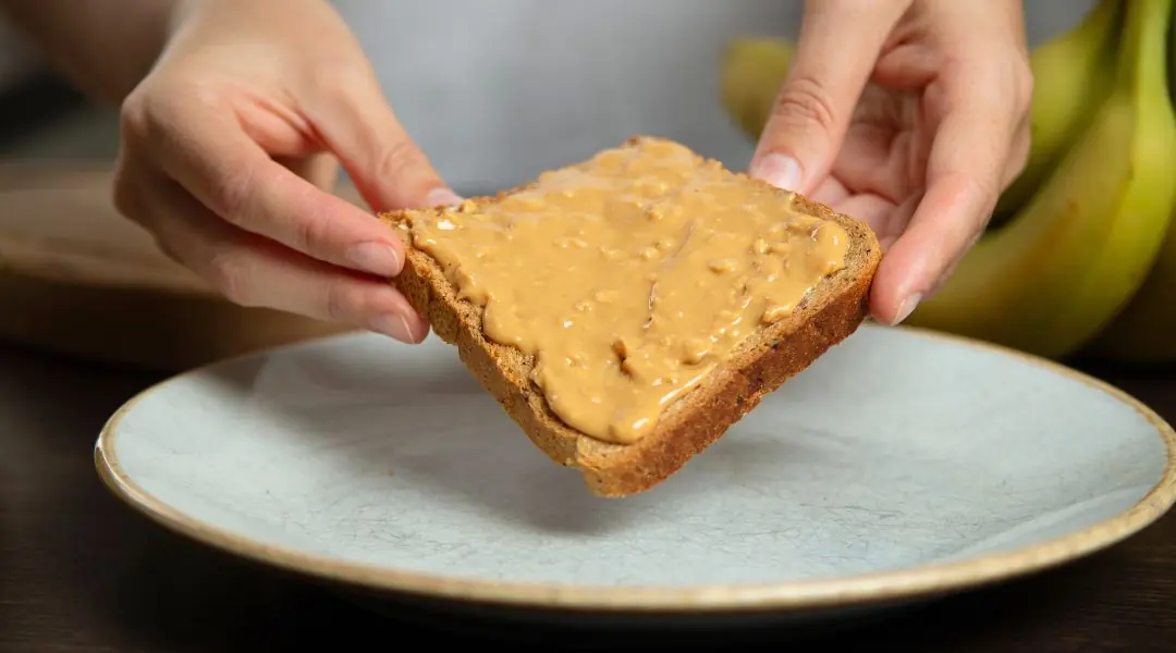 Hands holding a slice of bread with peanut butter over a plate, with bananas in the background
