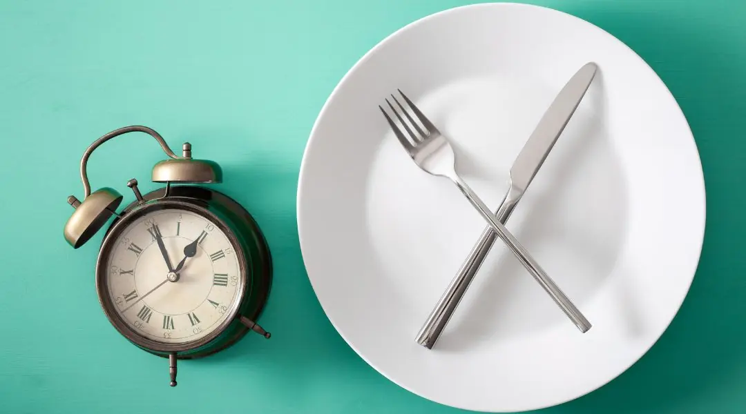 Assiette blanche vide avec une fourchette et un couteau croisés, à côté d'une horloge vintage sur un fond turquoise