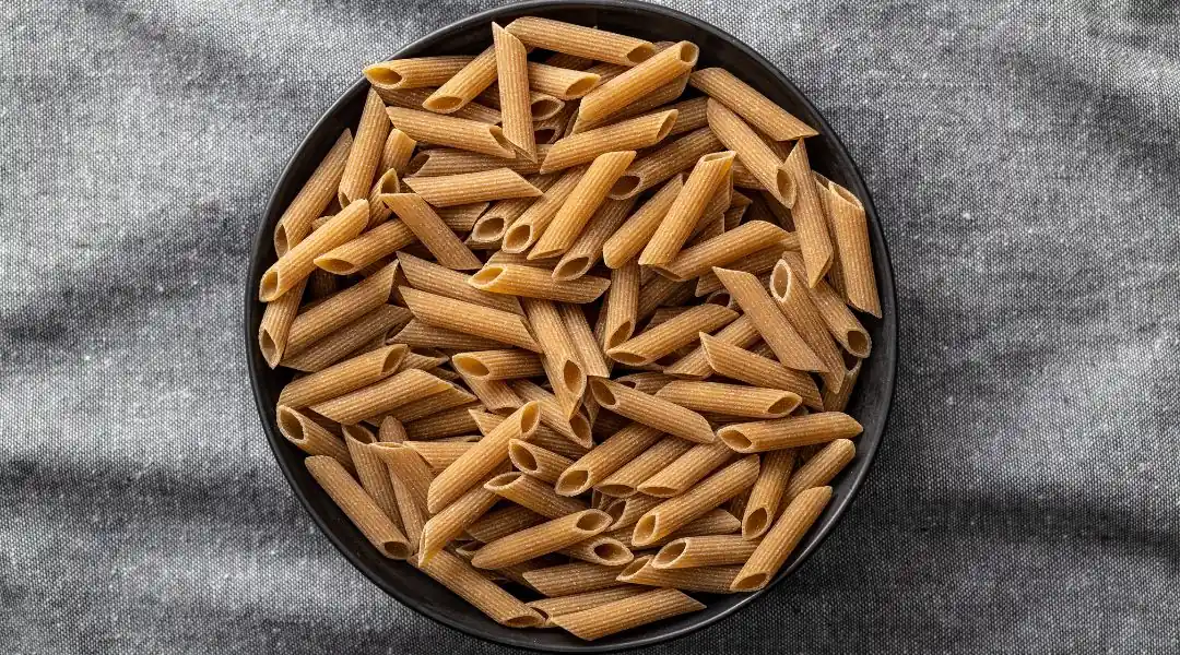 Pâtes penne à grains entiers crues dans un bol foncé sur tissu gris