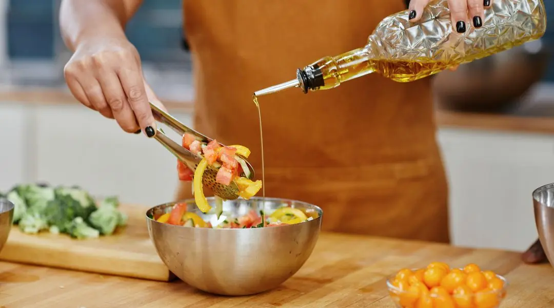 Mains versant de l'huile d'olive sur une salade colorée dans un bol, avec des légumes sur le plan de travail