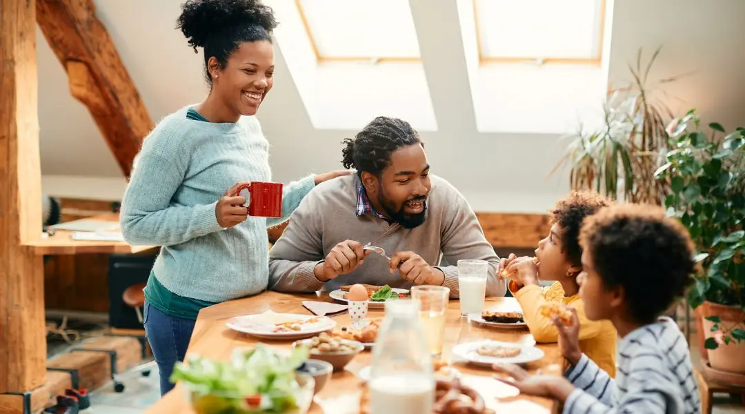 Famille souriante partageant un petit-déjeuner varié autour d'une table en bois dans une pièce lumineuse