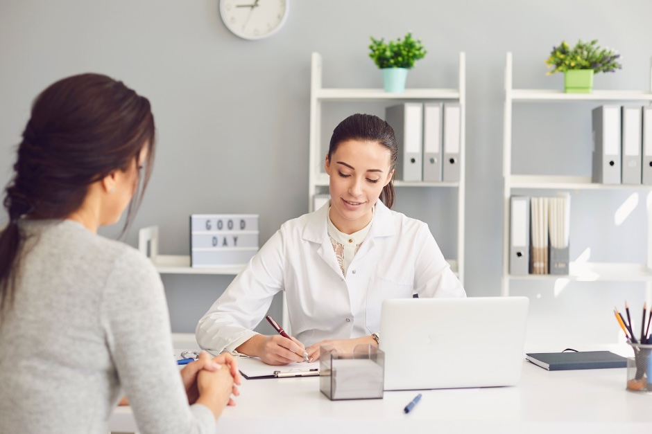 Une nutritionniste-diététiste en consultation avec une patiente à son bureau, prenant des notes en souriant, avec un ordinateur portable et des dossiers en arrière-plan. A female dietitian taking notes during a consultation with a patient in a modern office.