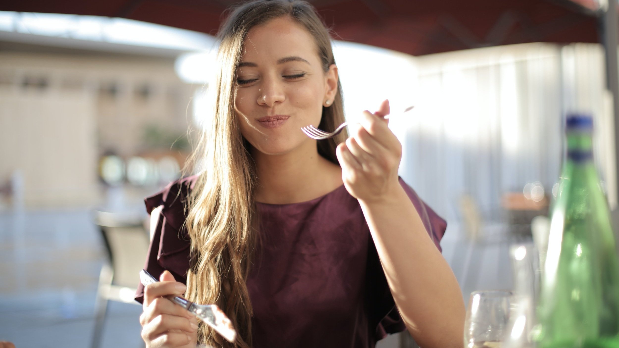 Une femme savourant un repas en extérieur, souriant les yeux fermés, tenant une fourchette dans une main. A woman enjoying a meal outdoors, smiling with her eyes closed while holding a fork in one hand.