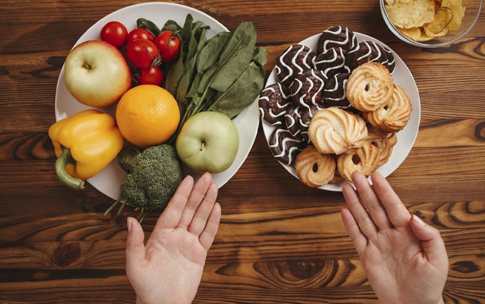 Deux assiettes sur une table en bois, l'une avec des fruits et légumes, l'autre avec des biscuits et pâtisseries, avec des mains désignant les deux. Two plates on a wooden table, one with fruits and vegetables, the other with cookies and pastries, with hands gesturing towards both.
