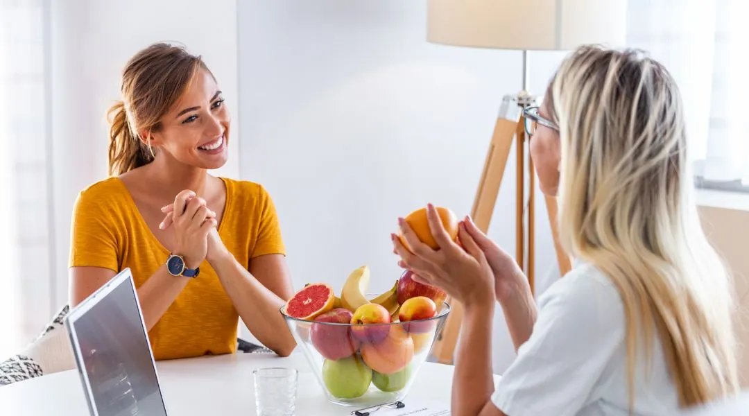 Nutirtionniste souriante en haut jaune discutant avec une autre femme près d’un bol de fruits frais
