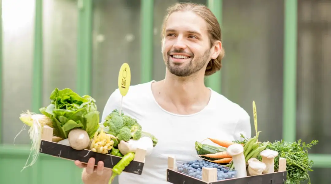 Homme souriant en t-shirt blanc tenant deux cagettes de légumes bio frais et de myrtilles