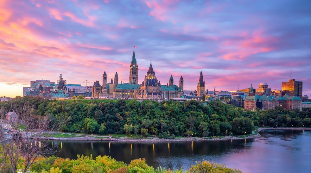 The Parliament Hill buildings in Ottawa, Canada, are silhouetted against a vibrant pink and purple sunset sky, with a river flowing in the foreground and lush green trees lining the banks.