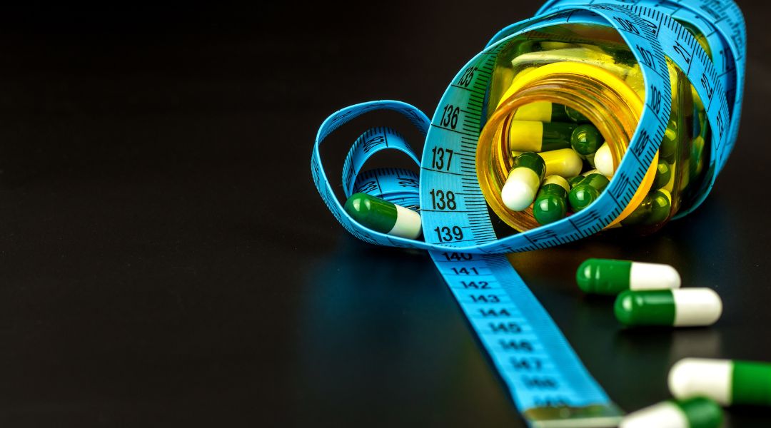 A blue measuring tape is wrapped around a bottle filled with green and white capsules, with some capsules scattered on a dark surface.