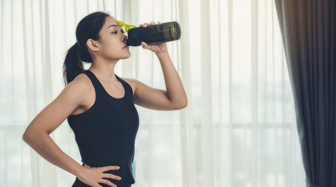 A person in athletic wear drinks from a dark water bottle with a yellow lid, standing with one hand on their hip.