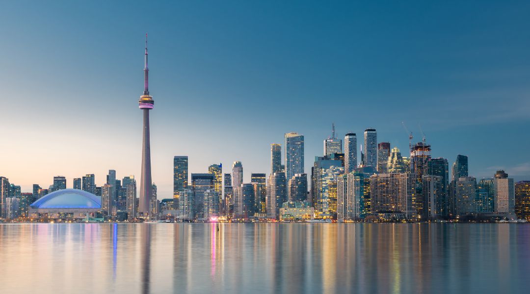 The Toronto skyline at dusk, featuring the CN Tower and illuminated buildings reflected in the water.