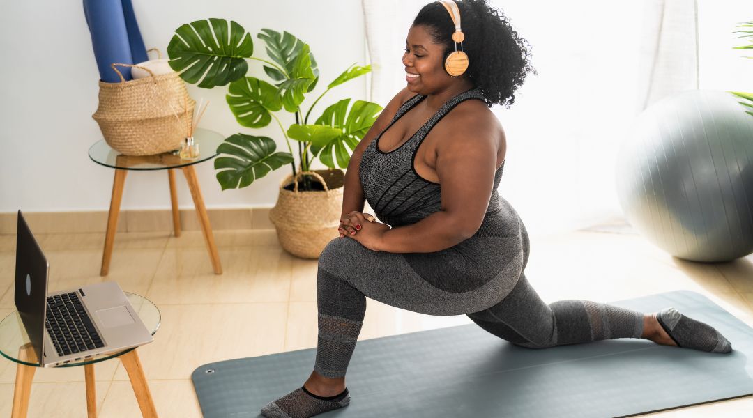 Une femme effectue des étirements sur un tapis de yoga dans son salon, avec un ordinateur portable à proximité pour suivre une séance en ligne.