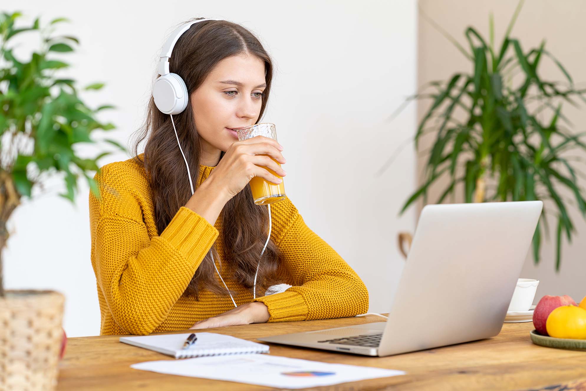 Woman wearing a yellow sweater and headphones, drinking orange juice while working on a laptop