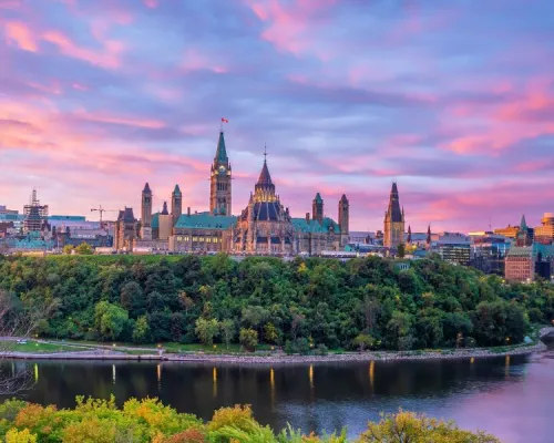 The Parliament Hill buildings in Ottawa, Canada, are silhouetted against a vibrant pink and purple sunset sky, with a river flowing in the foreground and lush green trees lining the banks.