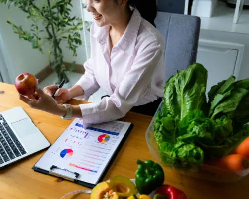 A person sitting at a table with a laptop, charts, a measuring tape, and a variety of fresh fruits and vegetables, including lettuce, carrots, bell peppers, and apples.