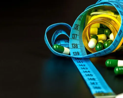 A blue measuring tape is wrapped around a bottle filled with green and white capsules, with some capsules scattered on a dark surface.