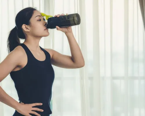 A person in athletic wear drinks from a dark water bottle with a yellow lid, standing with one hand on their hip.