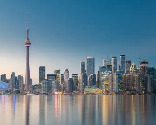 The Toronto skyline at dusk, featuring the CN Tower and illuminated buildings reflected in the water.