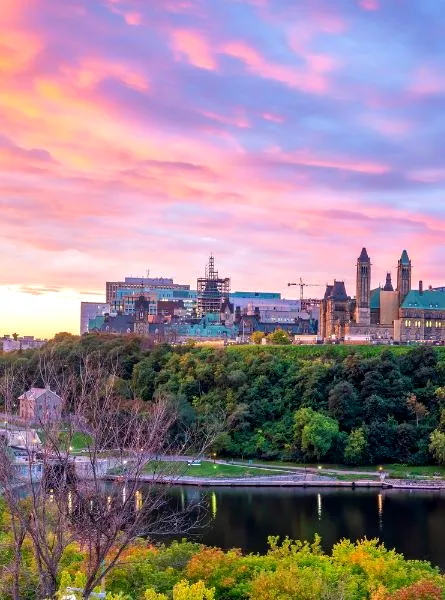 The Parliament Hill buildings in Ottawa, Canada, are silhouetted against a vibrant pink and purple sunset sky, with a river flowing in the foreground and lush green trees lining the banks.