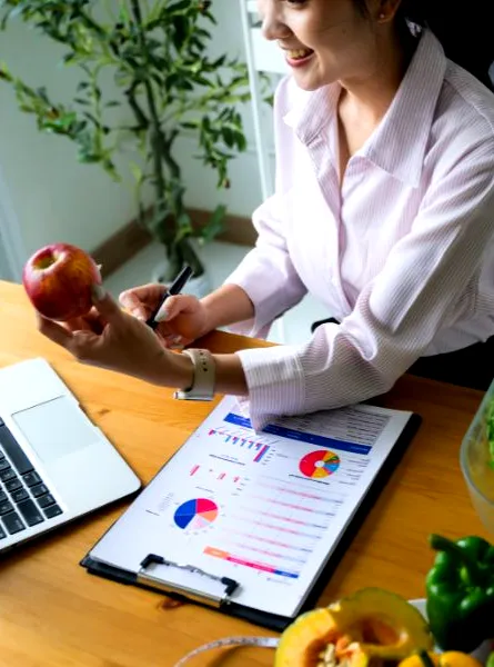 A person sitting at a table with a laptop, charts, a measuring tape, and a variety of fresh fruits and vegetables, including lettuce, carrots, bell peppers, and apples.