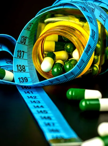 A blue measuring tape is wrapped around a bottle filled with green and white capsules, with some capsules scattered on a dark surface.