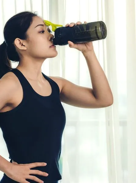 A person in athletic wear drinks from a dark water bottle with a yellow lid, standing with one hand on their hip.