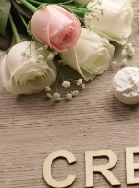 Cream and pink roses with small white blossoms arranged on a light wooden background next to a wooden spoon-shaped scoop and the word “CREATINE” in gold letters.