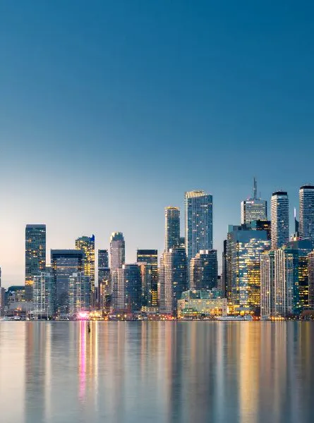 The Toronto skyline at dusk, featuring the CN Tower and illuminated buildings reflected in the water.